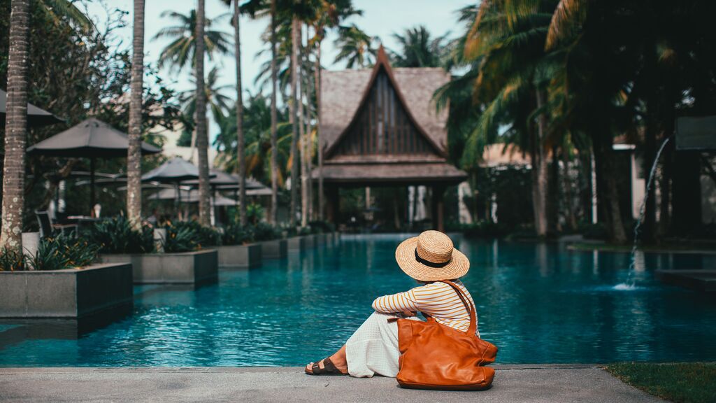 A guest relaxes beside a tranquil tropical resort pool surrounded by swaying palm trees and lush gardens.