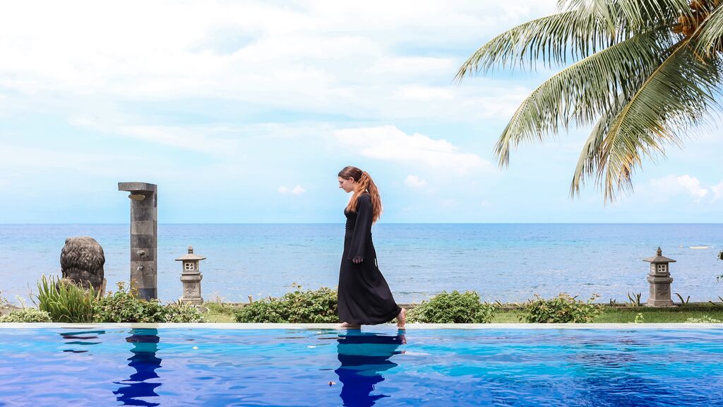 A guest strolls beside a beachfront infinity pool framed by swaying palm trees and panoramic ocean views.