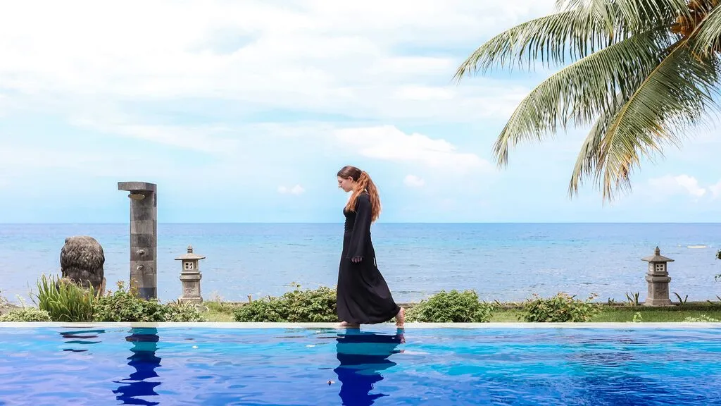 A guest strolls beside a beachfront infinity pool framed by swaying palm trees and panoramic ocean views.
