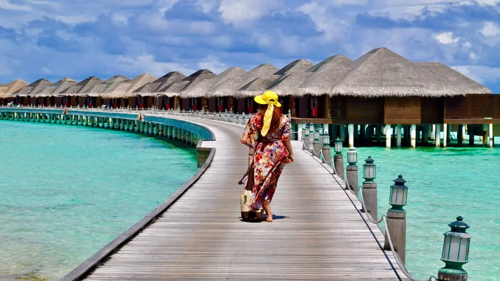 A guest walks along a wooden boardwalk toward overwater villas above crystal-clear turquoise waters.