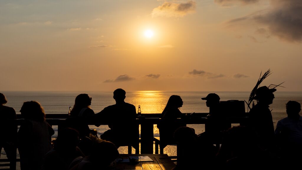 Silhouetted guests dining on an oceanfront terrace during a golden sunset.