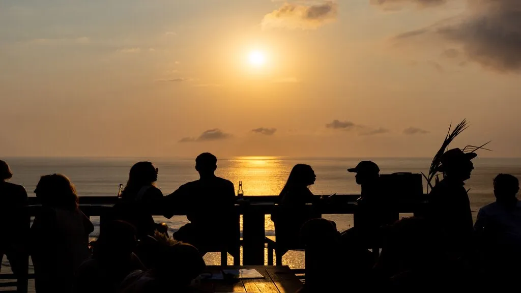 Silhouetted guests dining on an oceanfront terrace during a golden sunset.