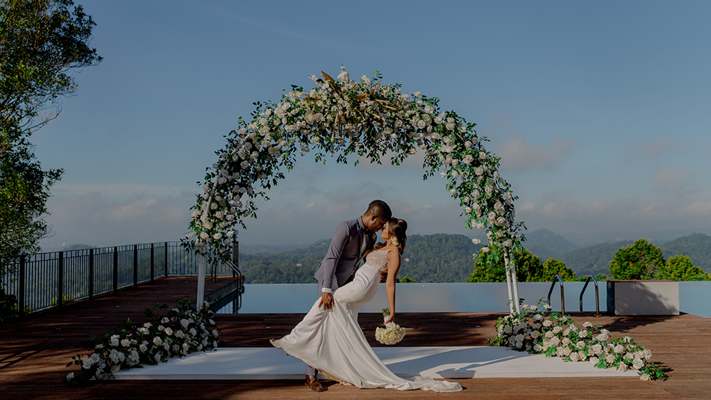 Bride and groom share a romantic moment beneath a floral arch overlooking an infinity pool and panoramic mountain views.