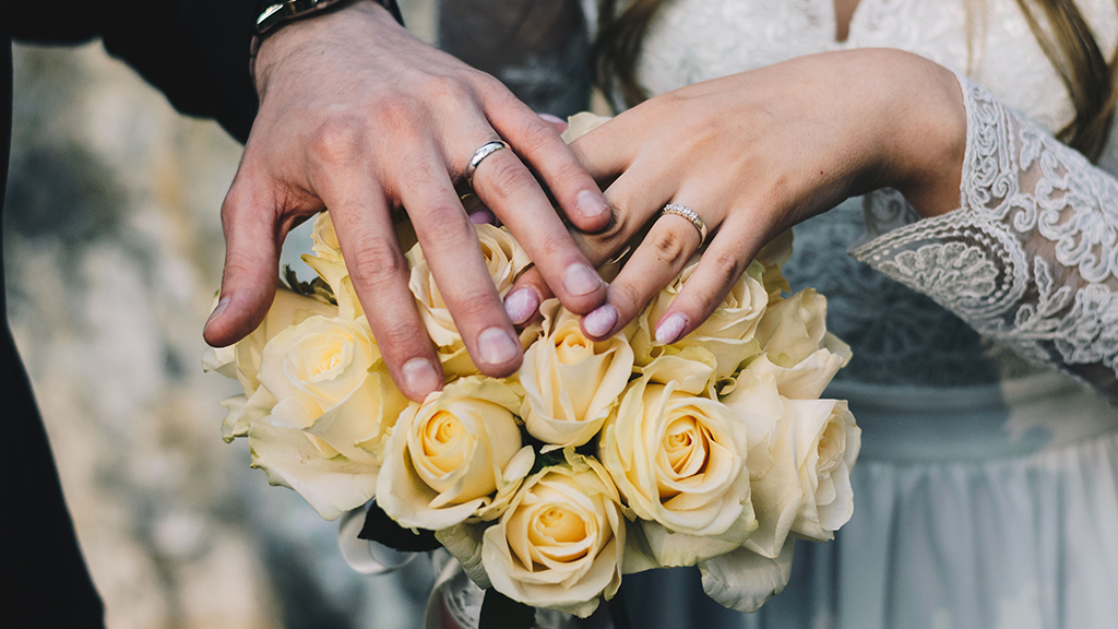 Close-up of a newlywed couple's hands with wedding rings resting on a bouquet of ivory roses.